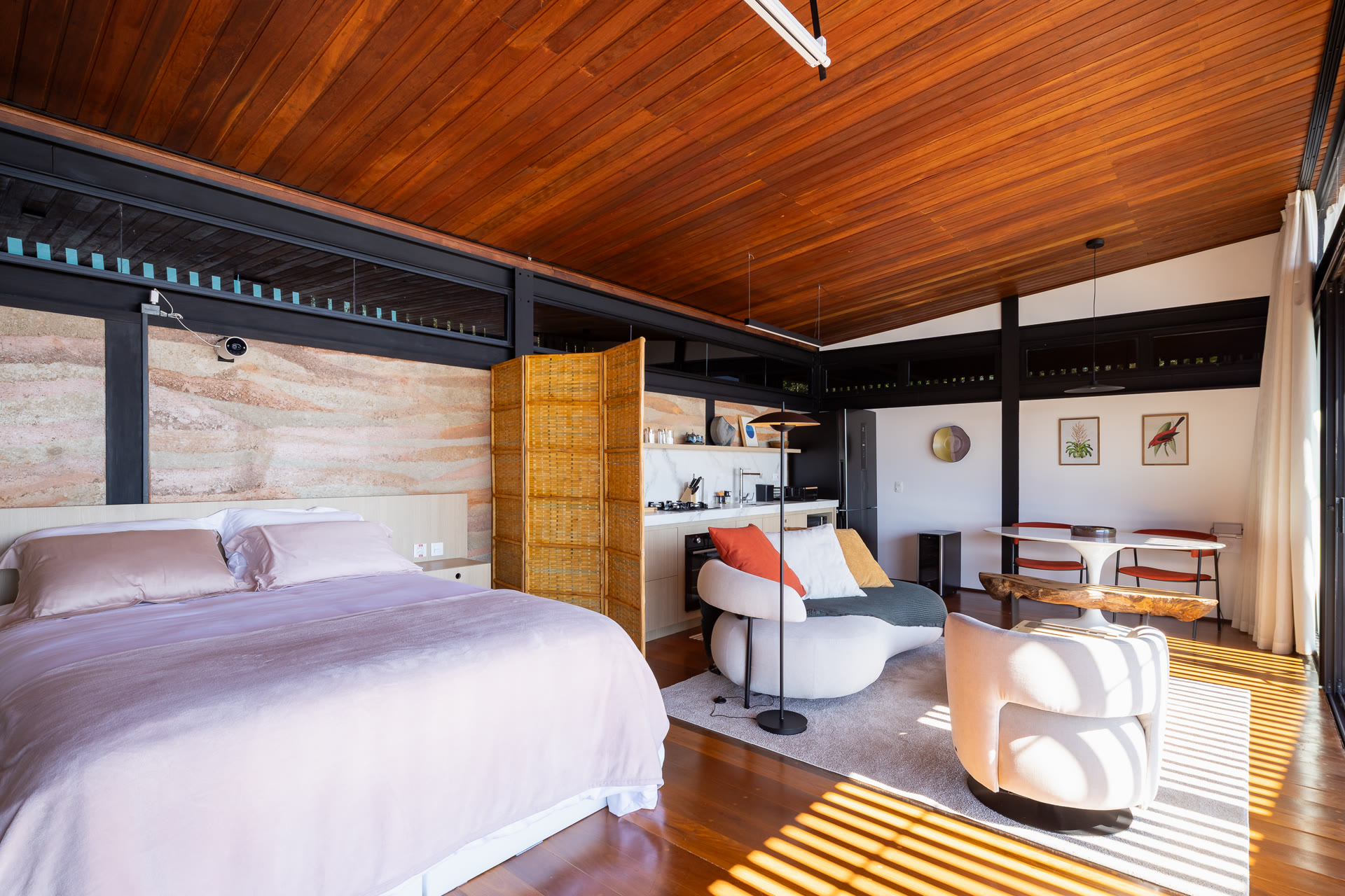 O interior do chalé: cama ampla, parede de taipa, biombo de palhinha e cozinha integrada sob o forro de madeira